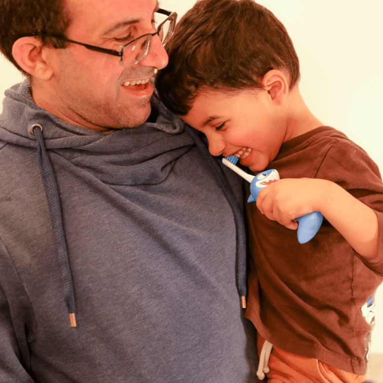 Young boy smiling and brushing teeth with shark toothbrush for kids while held by adult man